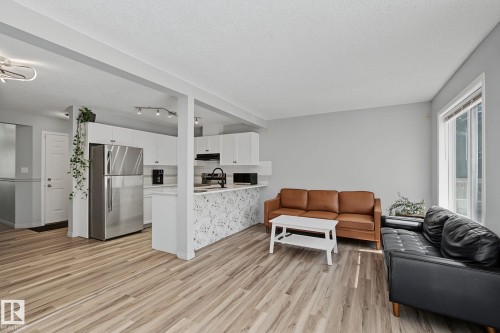 Open-concept living area featuring wood-finish flooring, a white kitchen with stainless steel appliances, and a kitchen island with a decorative panel - 5 14603 Miller Boulevard, Edmonton, AB 