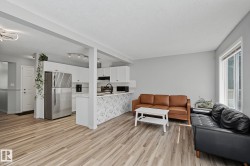 Open-concept living area featuring wood-finish flooring, a white kitchen with stainless steel appliances, and a kitchen island with a decorative panel - 