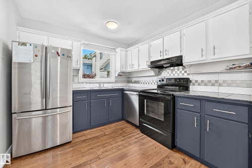 Kitchen featuring wood-finish flooring, two-tone cabinetry with white upper and blue lower cabinets, a checkerboard tile backsplash, and a window above the sink - 32 Mcleod Place, Edmonton, AB 