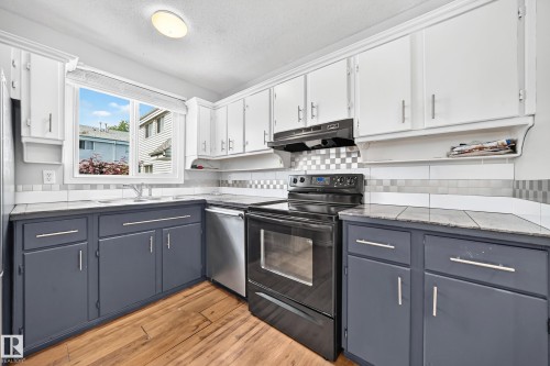 Kitchen featuring wood-finish flooring, two-tone cabinetry with white upper and blue-gray lower cabinets, a stainless steel dishwasher, and a black electric range with an overhead vent - 32 Mcleod Place, Edmonton, AB 