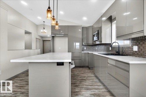 Kitchen featuring a large central island with a light-toned countertop, glossy gray cabinetry, a mosaic tile backsplash, wood-finish flooring, and pendant lighting fixtures - 1 Whitetail Way, Mundare, AB 
