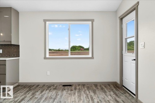 Contemporary kitchen area featuring grey cabinetry with a geometric tile backsplash, wood-finish flooring, a white exterior door with window panels, and a large window framing rural scenery - 1 Whitetail Way, Mundare, AB 