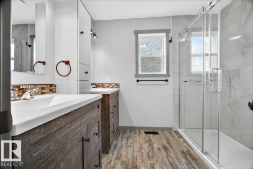 Spacious bathroom featuring a double vanity with wood-finish cabinetry, a tiled backsplash, a walk-in shower with a glass enclosure, and wood-finish flooring - 1 Whitetail Way, Mundare, AB 