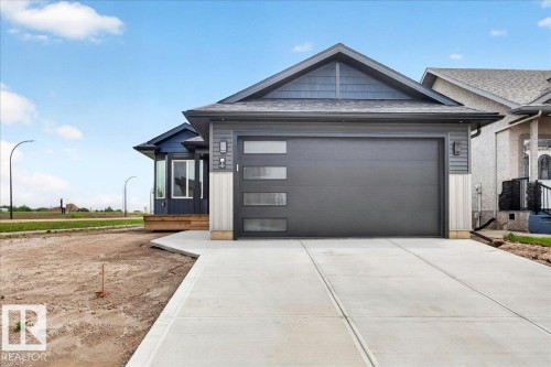 Contemporary exterior featuring dark gray siding with contrasting light vertical accents, a modern garage door with frosted glass panels, a new concrete driveway, and a covered front entry with a wood-finish porch - 1 Whitetail Way, Mundare, AB 