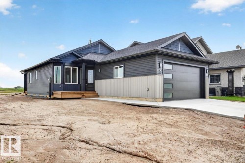 Contemporary exterior featuring dark gray and blue siding, a modern garage door with frosted glass panels, a concrete driveway, a front entry with a wood deck and steps, and multiple large windows - 1 Whitetail Way, Mundare, AB 