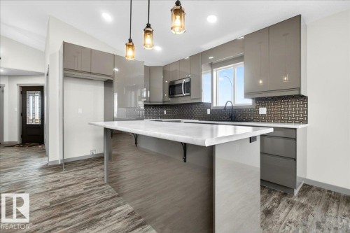 Contemporary kitchen featuring high-gloss gray cabinetry, a white countertop island with seating, a black patterned backsplash, and stainless steel appliances - 1 Whitetail Way, Mundare, AB 