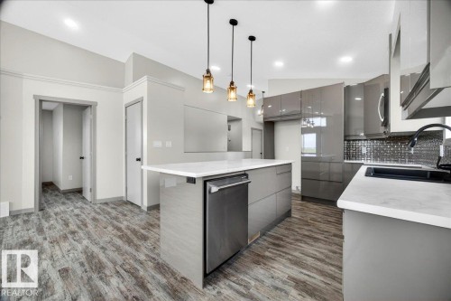 Contemporary kitchen featuring wood-finish flooring, a central island with a built-in dishwasher, pendant lighting, and a backsplash with reflective tiles - 1 Whitetail Way, Mundare, AB 