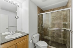 Three-piece bathroom featuring a glass-enclosed shower with stone-look tile surround and mosaic floor, a white toilet, and a vanity with a light wood-finish cabinet, speckled countertop, and white oval sink - 