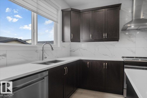 Modern kitchen featuring dark wood-finish cabinetry, white countertops, a stainless steel double sink with a gooseneck faucet, a white tile backsplash with subtle veining, and integrated stainless steel appliances - 2852 Duke Crescent, Edmonton, AB 