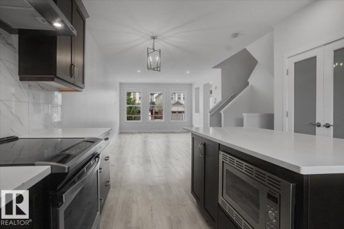 Open-concept living area featuring a kitchen island with built-in microwave, light wood-finish flooring, a contemporary chandelier, multiple windows, and a staircase with white railings - 2852 Duke Crescent, Edmonton, AB 