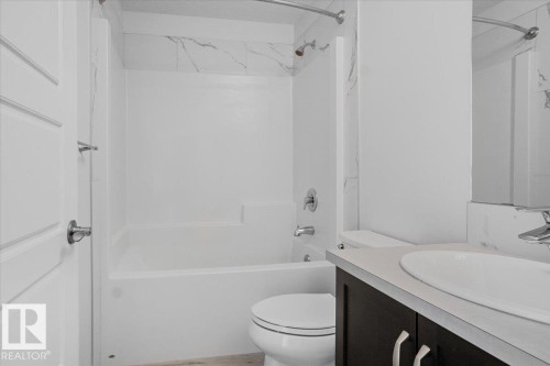 Bathroom featuring a white tub/shower combination with marble-patterned tile surround, a dark wood-finish vanity with a light countertop, and a white toilet - 2852 Duke Crescent, Edmonton, AB 
