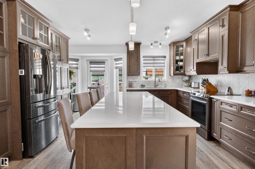 Gourmet kitchen featuring an oversized central island with seating, stainless steel appliances, extensive cabinetry with glass-front accents, light-toned countertops, and a full-height herringbone backsplash - 5006 53 Avenue, Cold Lake, AB 