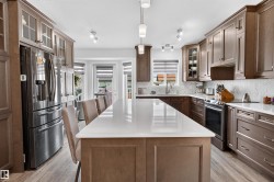 Gourmet kitchen featuring an oversized central island with seating, stainless steel appliances, extensive cabinetry with glass-front accents, light-toned countertops, and a full-height herringbone backsplash - 