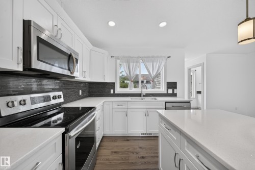 Modern kitchen featuring white cabinetry, stainless steel appliances, dark tile backsplash, a center island, and wood-finish flooring - 227 Cy Becker Blvd, Edmonton, AB 
