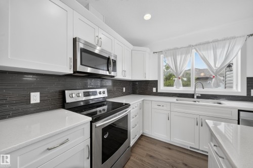 Kitchen featuring white shaker-style cabinetry, stainless steel appliances, dark linear tile backsplash, light-toned countertops, and wood-finish flooring - 227 Cy Becker Blvd, Edmonton, AB 