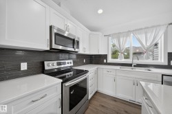 Kitchen featuring white shaker-style cabinetry, stainless steel appliances, dark linear tile backsplash, light-toned countertops, and wood-finish flooring - 