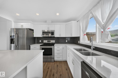 Kitchen featuring white shaker cabinetry, stainless steel appliances, dark tile backsplash, light-toned countertops, and wood-finish flooring - 227 Cy Becker Blvd, Edmonton, AB 