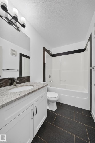 Bathroom featuring white cabinetry, a single basin sink, a speckled countertop, a shower-tub combination, and dark rectangular floor tiles - 227 Cy Becker Blvd, Edmonton, AB 