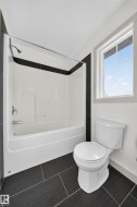 Bathroom featuring a white bathtub and shower combination with chrome fixtures, a two-piece toilet, dark large-format floor tiles, and a white framed window - 