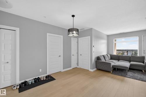 Open-concept living area featuring light wood-finish flooring, a contemporary ceiling light fixture, a large window offering expansive outdoor scenery, and neutral gray wall paint - 706 5151 Windermere Boulevard, Edmonton, AB 