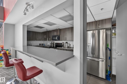 Contemporary kitchen with a pass-through bar, featuring dark wood-finish cabinetry, stainless steel appliances, and a gray countertop - 706 5151 Windermere Boulevard, Edmonton, AB 