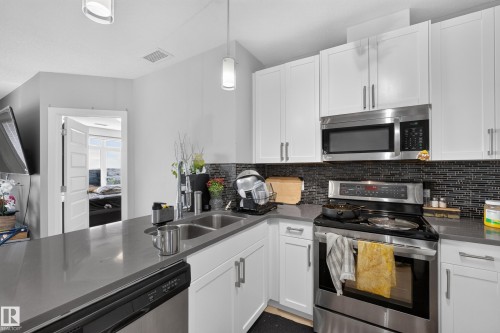 Modern kitchen featuring white shaker cabinetry, stainless steel appliances, a dark tile backsplash, and light gray countertops - 706 5151 Windermere Boulevard, Edmonton, AB 