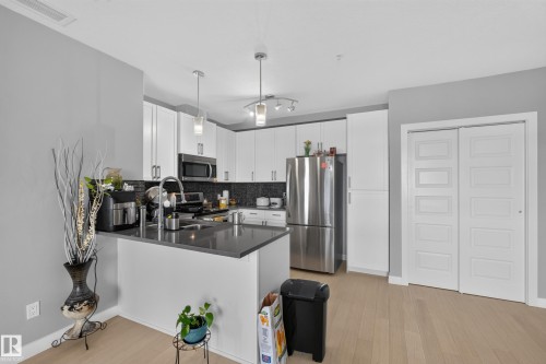 Modern kitchen featuring white cabinetry, stainless steel appliances, a dark countertop, a dark patterned backsplash, and wood-finish flooring - 706 5151 Windermere Boulevard, Edmonton, AB 