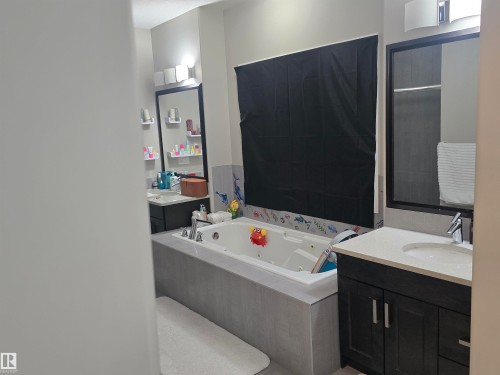 Bathroom featuring a built-in jetted tub with tile surround, dual vanity areas with dark wood cabinetry and light countertops, and individual framed mirrors with integrated lighting - 1019 175 Street, Edmonton, AB 