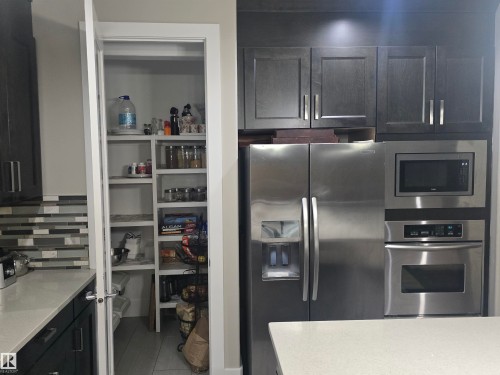 Kitchen featuring stainless steel refrigerator, built-in microwave, wall oven, dark wood cabinetry, and a walk-in pantry with extensive shelving - 1019 175 Street, Edmonton, AB 