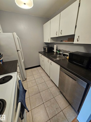 Galley kitchen featuring white cabinetry, dark countertops, a stainless steel dishwasher, a single basin sink, and tile flooring - 10838 108 Street Nw, Edmonton, AB 