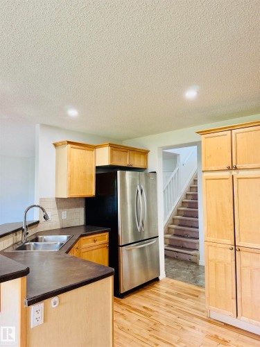 Kitchen featuring wood-finish flooring, light wood cabinetry, dark countertops, a stainless steel refrigerator, and a double basin sink with a gooseneck faucet - 3120 25 Avenue, Edmonton, AB 