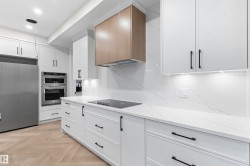 White cabinetry with matte black hardware, integrated stainless steel appliances, wood-finish flooring in a herringbone pattern, wood-finish range hood, and white countertops with a matching backsplash - 