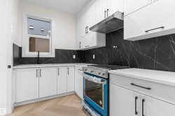 Modern kitchen featuring white cabinetry, black stone backsplash with white veining, light wood-finish herringbone flooring, stainless steel range, and undermount sink with a black faucet - 