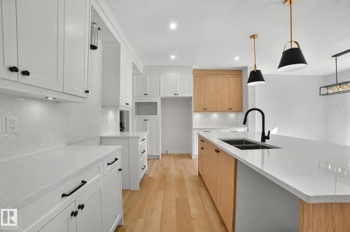 Spacious kitchen featuring an expansive island with a matte black faucet and dual basin sink, white shaker-style cabinetry, light wood-finish flooring, white countertops, and contemporary pendant lighting - 204 Falcon Crescent, Leduc, AB 