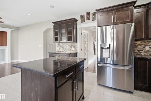 Kitchen featuring a central island with a dark stone countertop, dark wood cabinetry with glass-front uppers, a mosaic tile backsplash, and a stainless steel French door refrigerator - 16807 54 Street, Edmonton, AB - Indoor Photo Showing Kitchen With Stainless Steel Kitchen