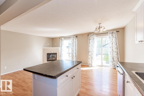 Kitchen island with dark countertop and white cabinetry, wood-finish flooring, built-in fireplace, and sliding glass doors providing access to the exterior - 39 Vega Avenue, Spruce Grove, AB - Indoor Photo Showing Kitchen With Fireplace