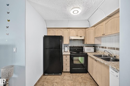 Compact kitchen featuring light wood cabinetry, a double basin stainless steel sink, black appliances, and a tiled backsplash - #208 9619 174 St Nw, Edmonton, AB 