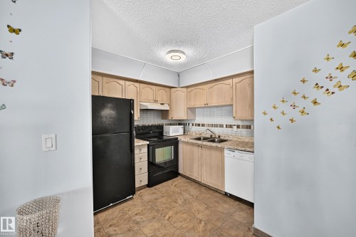 Kitchen featuring light-toned cabinetry, a double basin stainless steel sink, black range, black refrigerator, and an integrated white dishwasher - #208 9619 174 St Nw, Edmonton, AB 