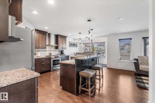 Open-concept kitchen featuring wood-finish flooring, dark wood cabinetry, granite-look countertops, stainless steel appliances, and a central island with bar seating - 6 Sedona Place, Fort Saskatchewan, AB - Indoor Photo Showing Kitchen With Upgraded Kitchen