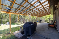 Expansive concrete patio featuring a clear panel roof with exposed wood framing, exterior wall-mounted lighting fixtures, and a natural tree-lined backdrop - 
