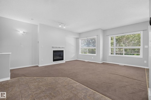 Living area featuring a built-in fireplace with a white mantel, two large windows, brown carpeting, and a tiled entry - #113 3010 33 Av Nw, Edmonton, AB 