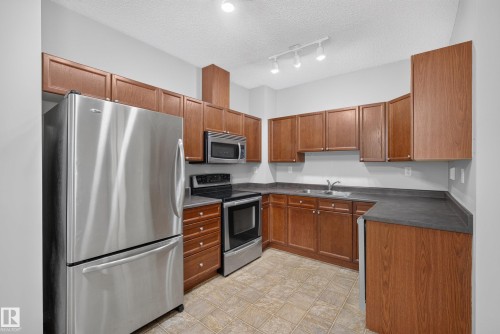 U-shaped kitchen featuring wood-finish cabinetry, dark countertops, and stainless steel appliances including a refrigerator, range, and microwave - #113 3010 33 Av Nw, Edmonton, AB 
