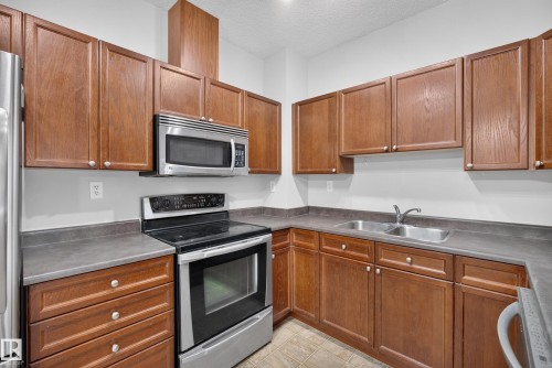 Kitchen featuring wood cabinetry, stainless steel appliances, a double basin sink, and a range with an overhead microwave - #113 3010 33 Av Nw, Edmonton, AB 