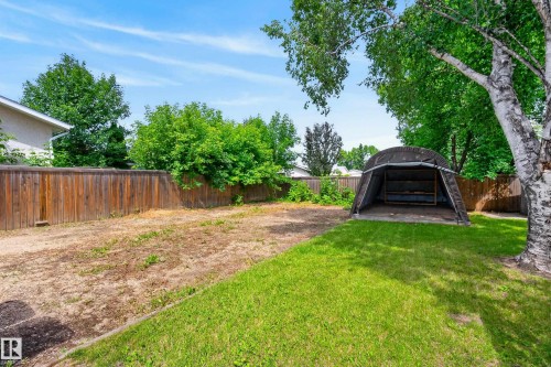 Expansive backyard featuring a partial manicured lawn, a large wood privacy fence, mature trees, and a fabric-covered shelter with a concrete base - 3 Bristow Crescent, Spruce Grove, AB - Outdoor