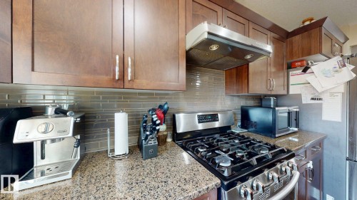 Kitchen featuring rich wood cabinetry, granular stone countertops, stainless steel gas range, coordinating range hood, and a tiled backsplash - 6824 Evans Wynd Nw, Edmonton, AB - Indoor Photo Showing Kitchen