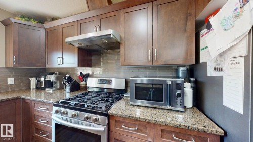 Kitchen featuring wood cabinetry, granite-style countertops, a stainless steel gas range with a coordinating range hood, and a tiled backsplash - 6824 Evans Wynd Nw, Edmonton, AB - Indoor Photo Showing Kitchen With Upgraded Kitchen