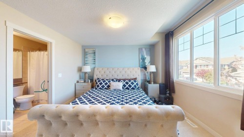 Spacious bedroom featuring large windows, light-colored walls, a ceiling-mounted light fixture, carpet flooring, and an open doorway revealing a partial bathroom with a toilet and shower - 6824 Evans Wynd Nw, Edmonton, AB - Indoor Photo Showing Bedroom
