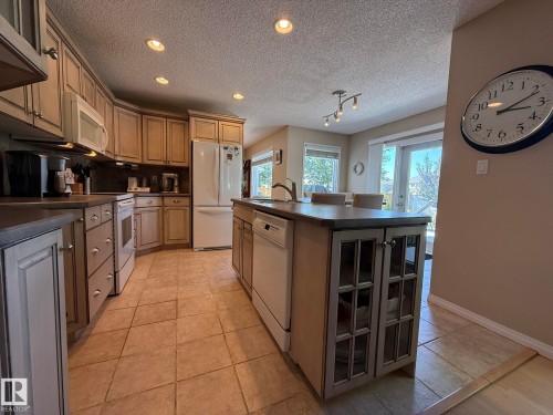 Open-concept kitchen featuring light-toned cabinetry, a central island with glass-front doors, recessed lighting, tile flooring, and integrated white appliances - 5604 57 Avenue, Beaumont, AB 