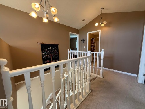 Upper level hallway featuring vaulted ceilings, neutral-toned carpeting, white painted balusters and handrails, and two multi-light pendant fixtures - 5604 57 Avenue, Beaumont, AB 