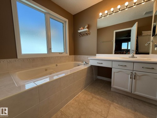 Built-in jetted tub with tiled surround, white dual-pane window, white vanity with integrated sink, large wall-mounted mirror, and tile flooring - 5604 57 Avenue, Beaumont, AB 